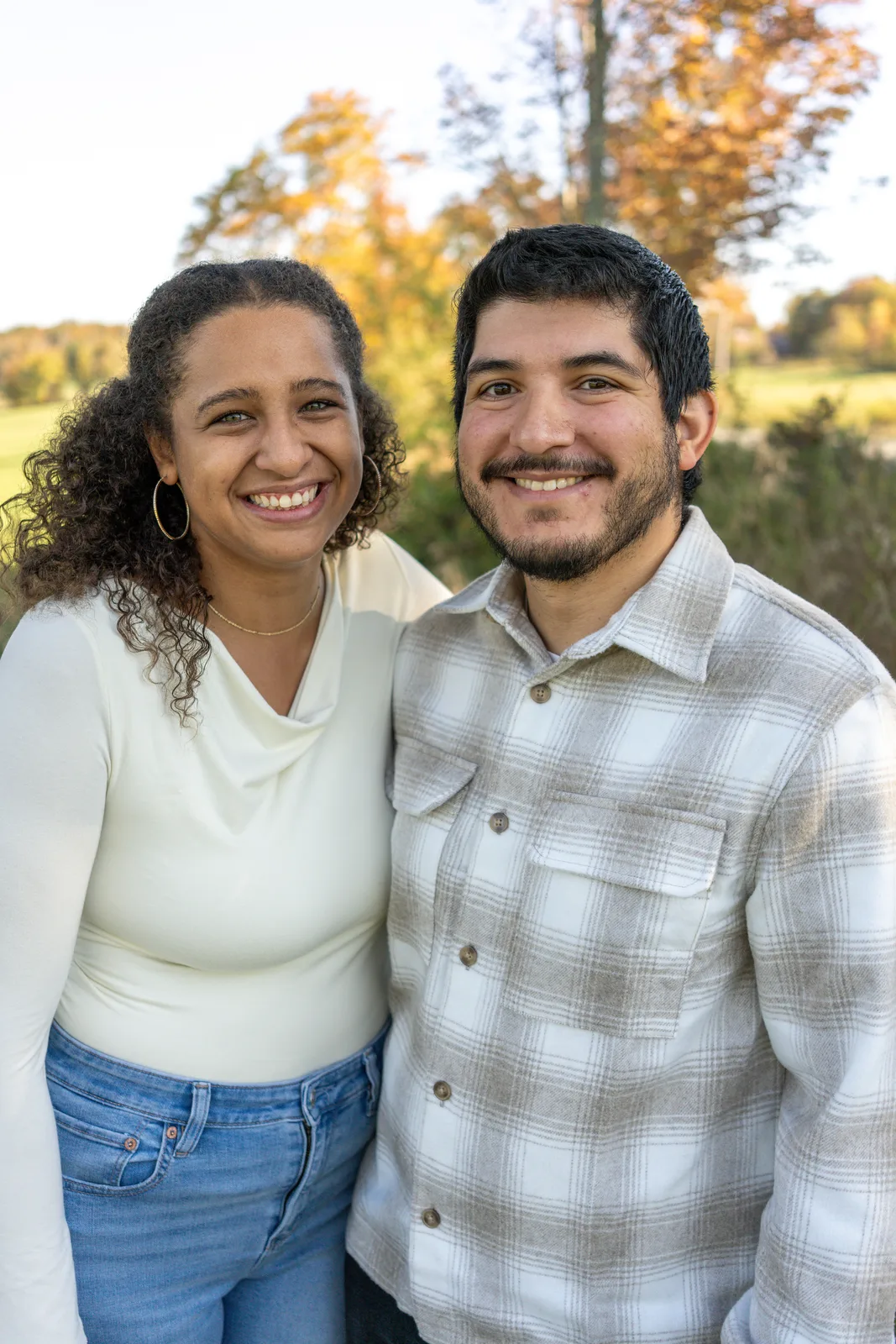 Two adults standing close in a field with autumn trees and open sky