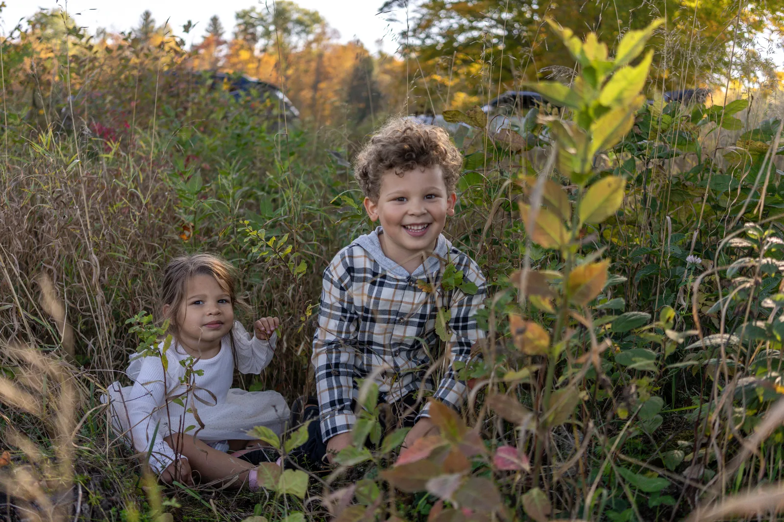 Two children in plaid and white clothing crouching in tall grass with autumn foliage behind