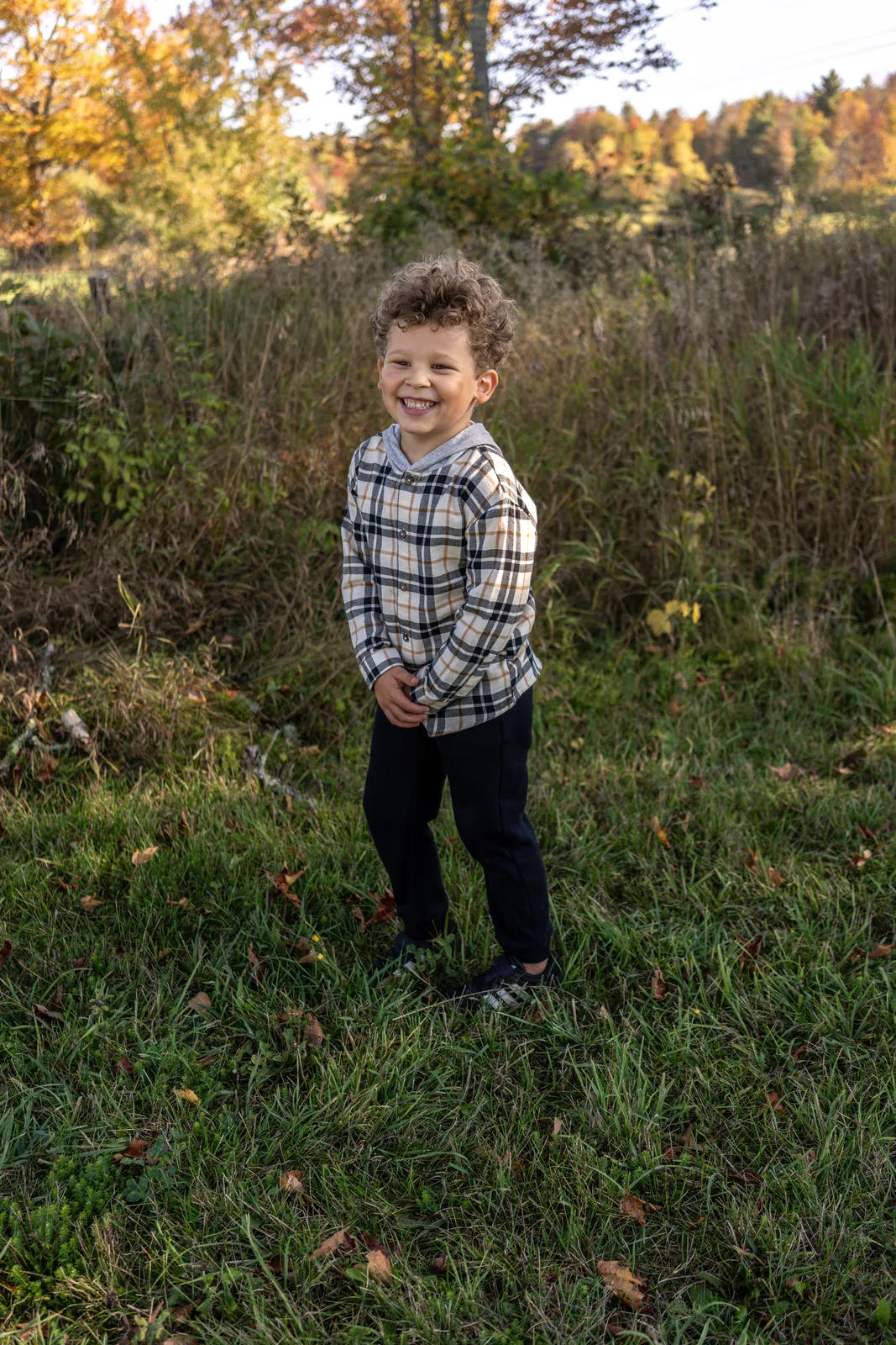 Child in plaid shirt and dark pants standing in a grassy field with autumn trees