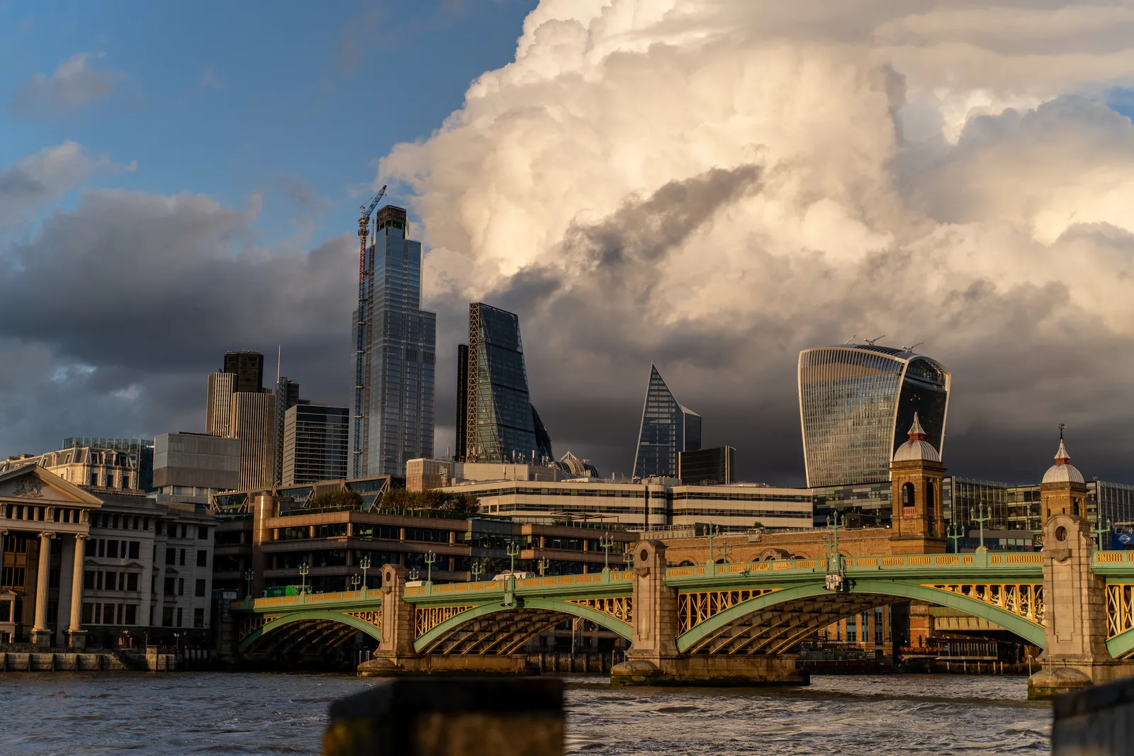 London skyline and a green-arched bridge across the River Thames beneath sunlit storm clouds