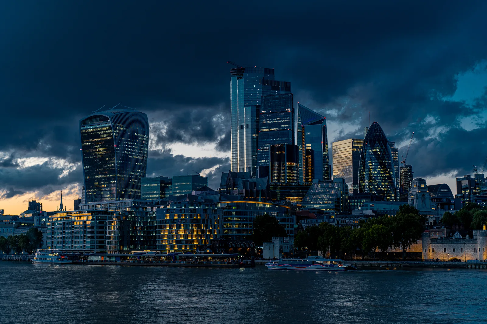 London office towers lit at dusk across the River Thames beneath heavy clouds