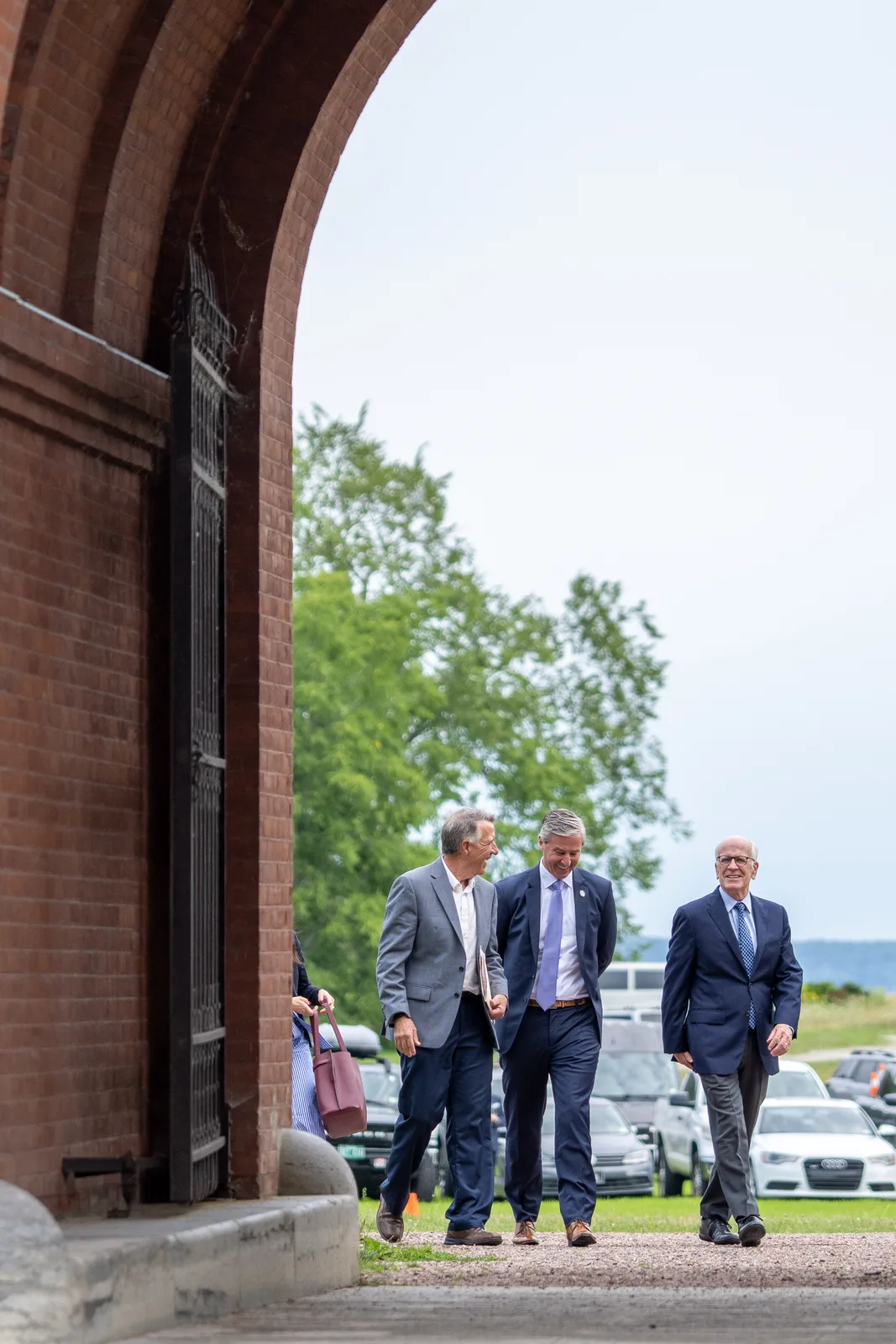 Three delegates approach through a brick arch, with the Shelburne Farms landscape beyond them.