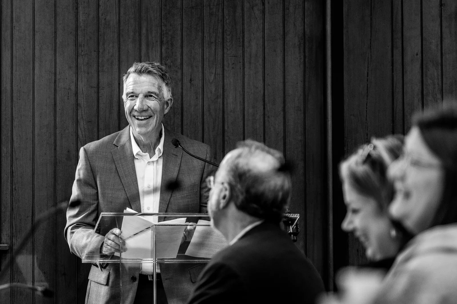 A smiling speaker addresses the room at a microphone in a black-and-white photograph.