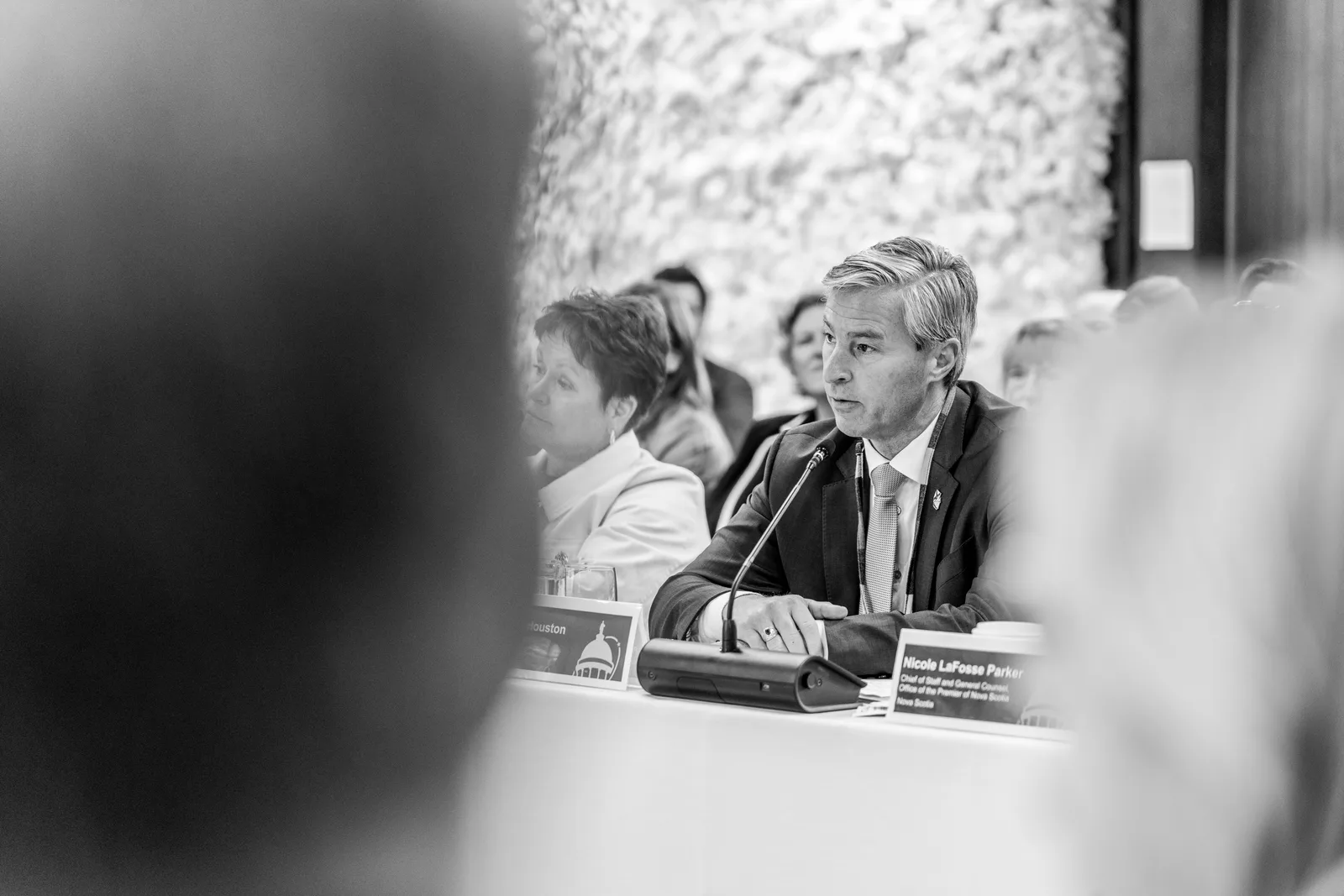 A speaker is framed between blurred attendees in a black-and-white view across the table.