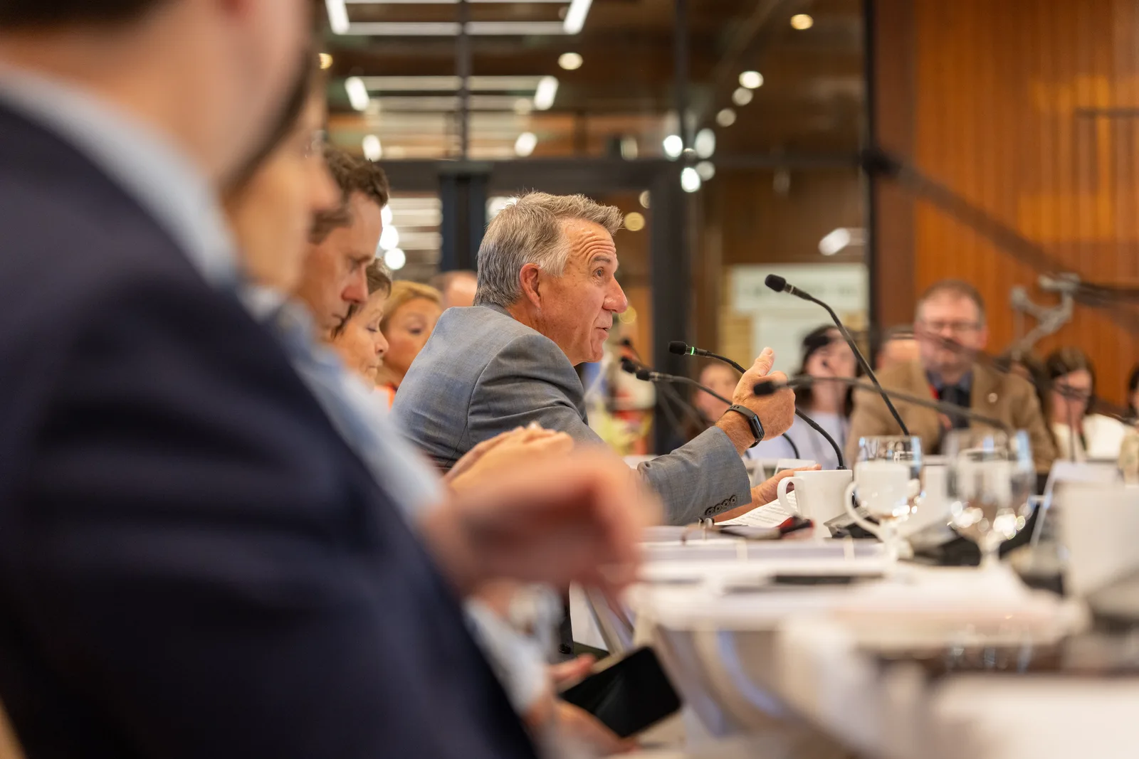 A delegate speaks across the hollow-square table as microphones and water glasses frame the foreground.