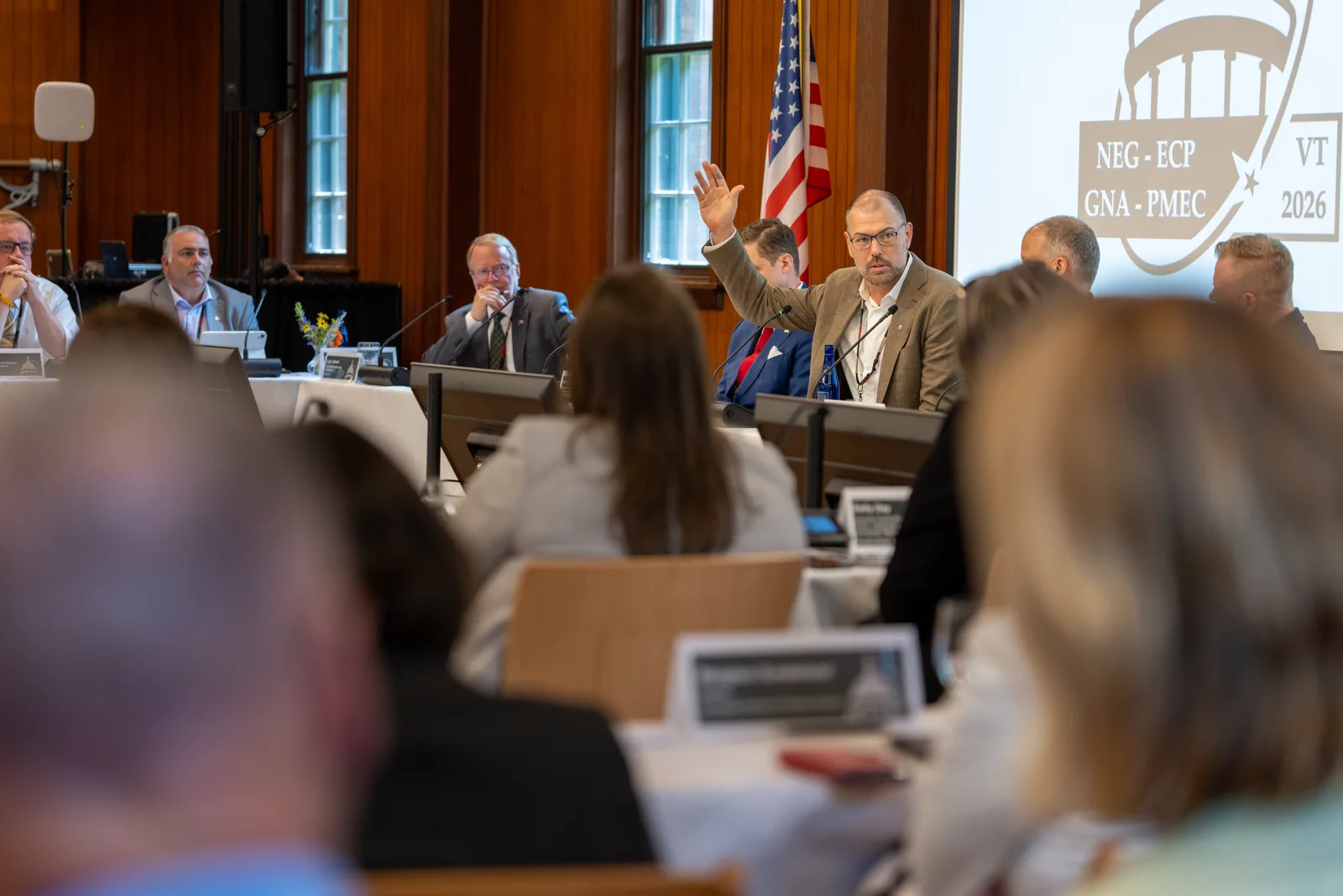 A summit delegate gestures while speaking beneath United States and Canadian flags inside the Coach Barn.