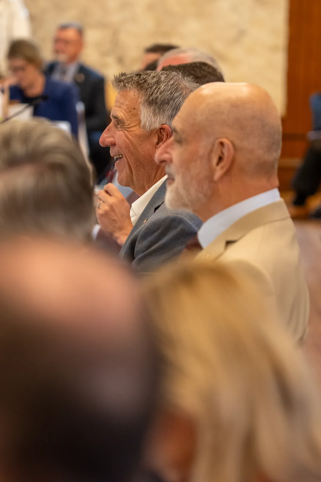Two gray-haired delegates talk across the table as other participants blur into the foreground.