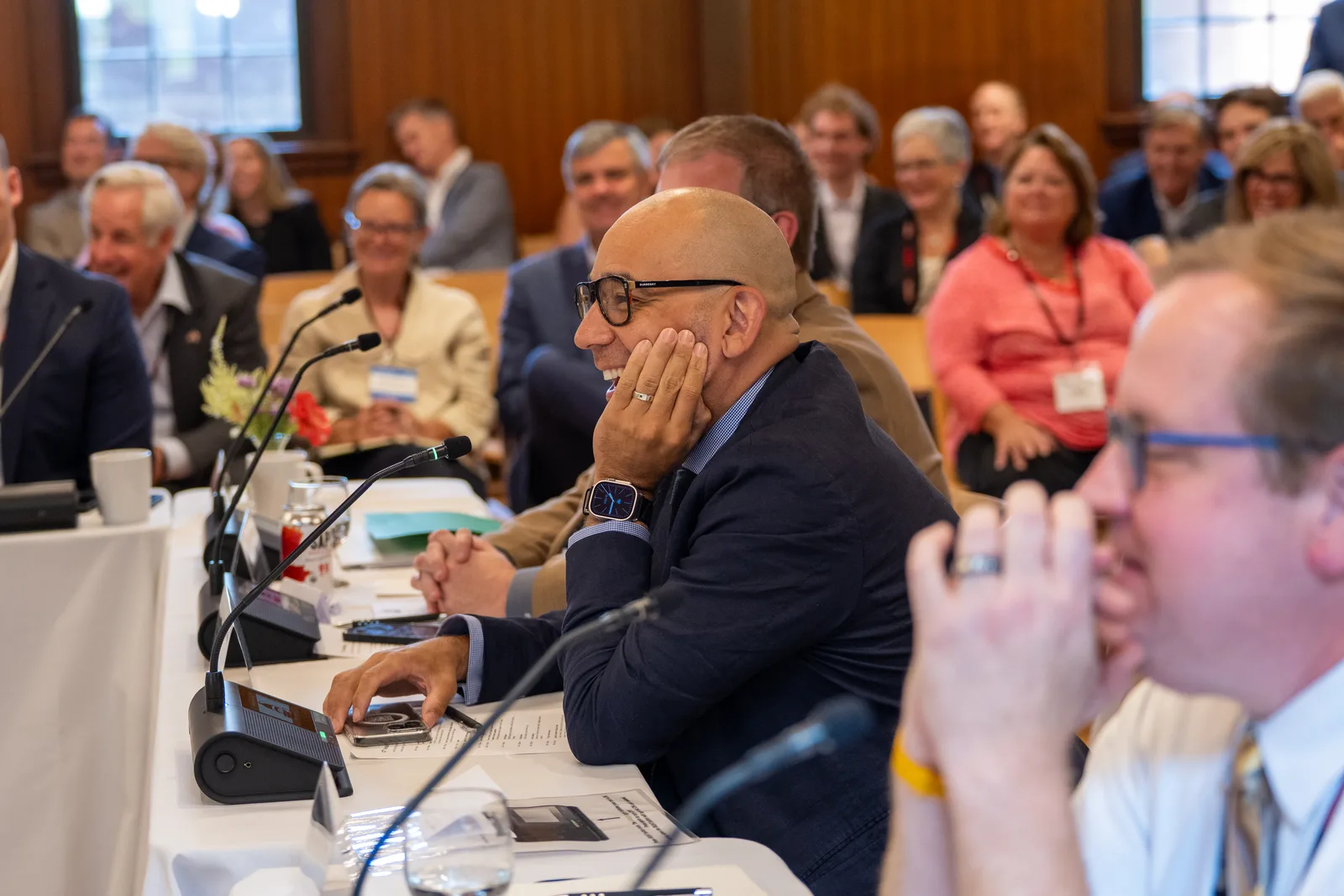 A delegate wearing glasses listens at the table with a full audience seated behind him.