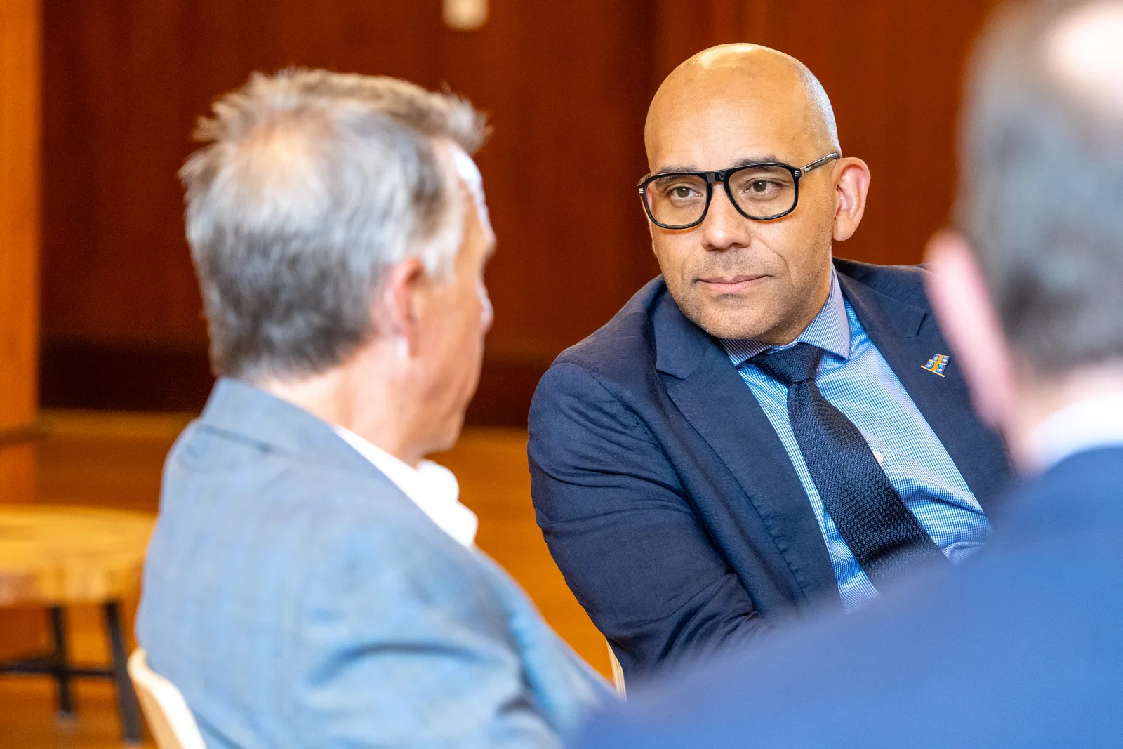 A delegate wearing black glasses listens as another participant speaks across the table.