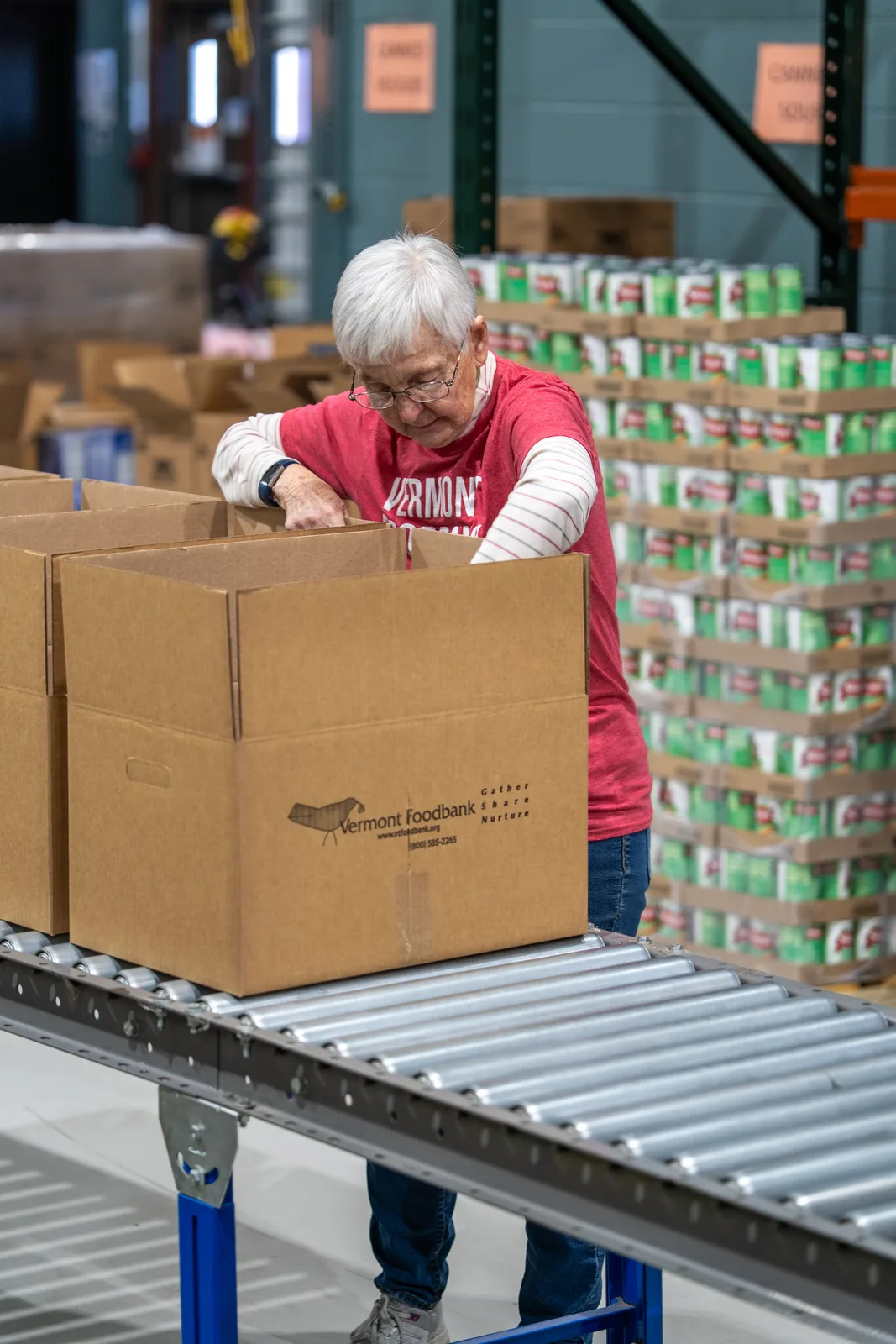 Person in red shirt packing cardboard boxes on a conveyor in a warehouse with stacked cans