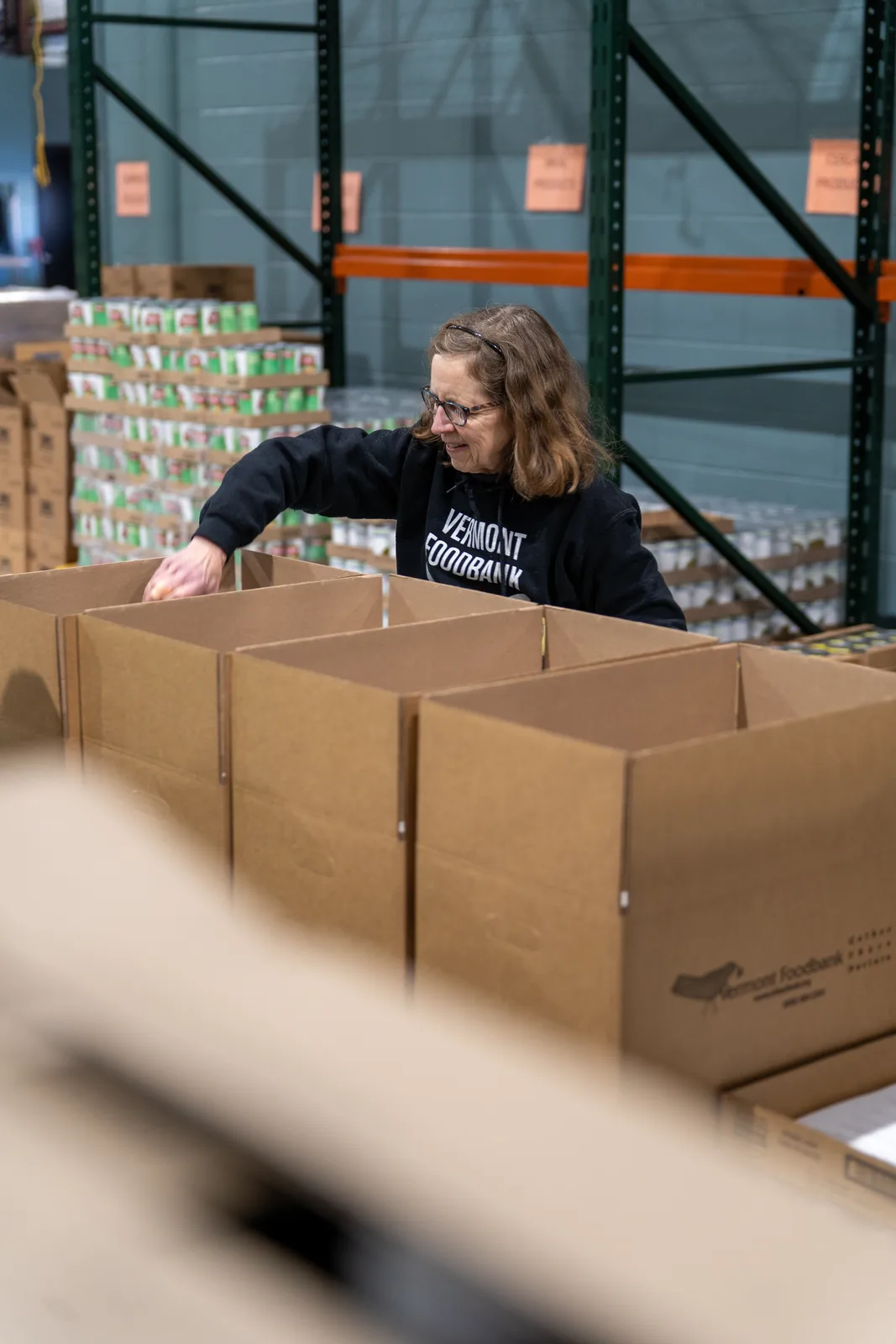 Person in black sweatshirt arranging empty cardboard boxes in a warehouse with shelves of cans