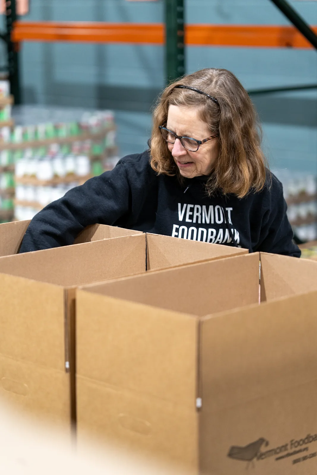 Person in black sweatshirt leaning over open cardboard boxes in a warehouse with shelves
