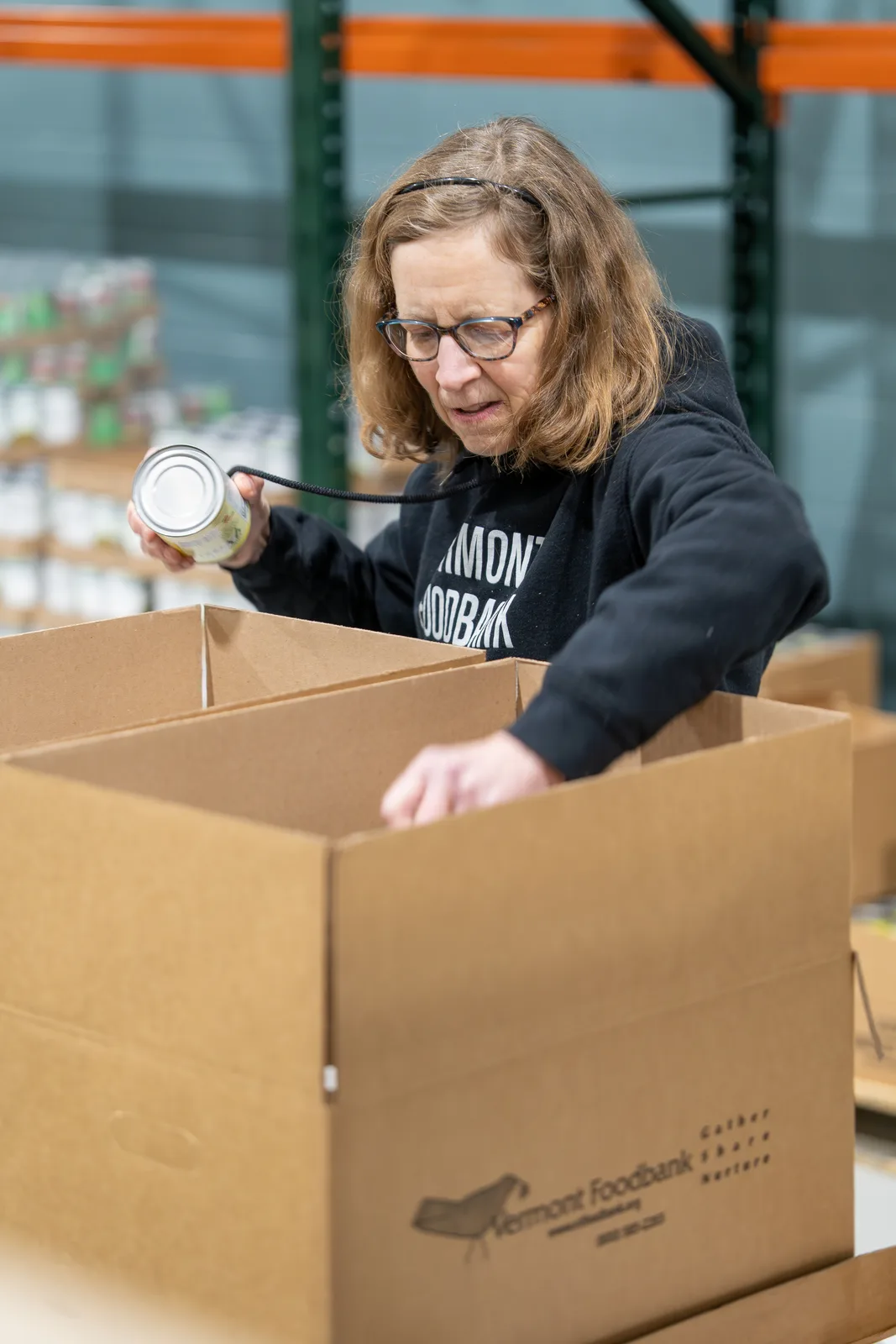 Person in black sweatshirt placing a can into an open cardboard box in a warehouse