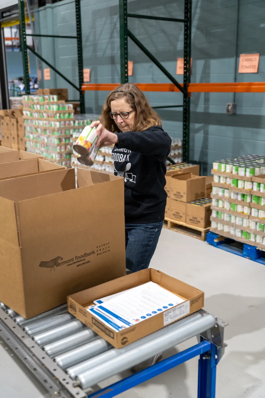 Person in black sweatshirt pouring canned food into a box on a conveyor in a warehouse