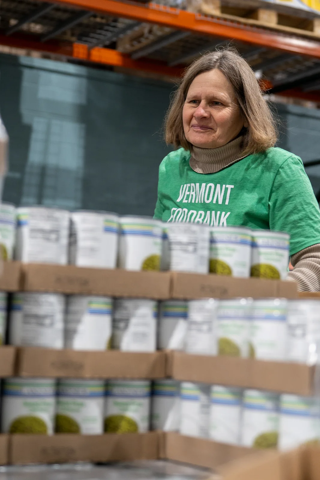 Person in green shirt standing behind stacked boxes of canned goods in a warehouse