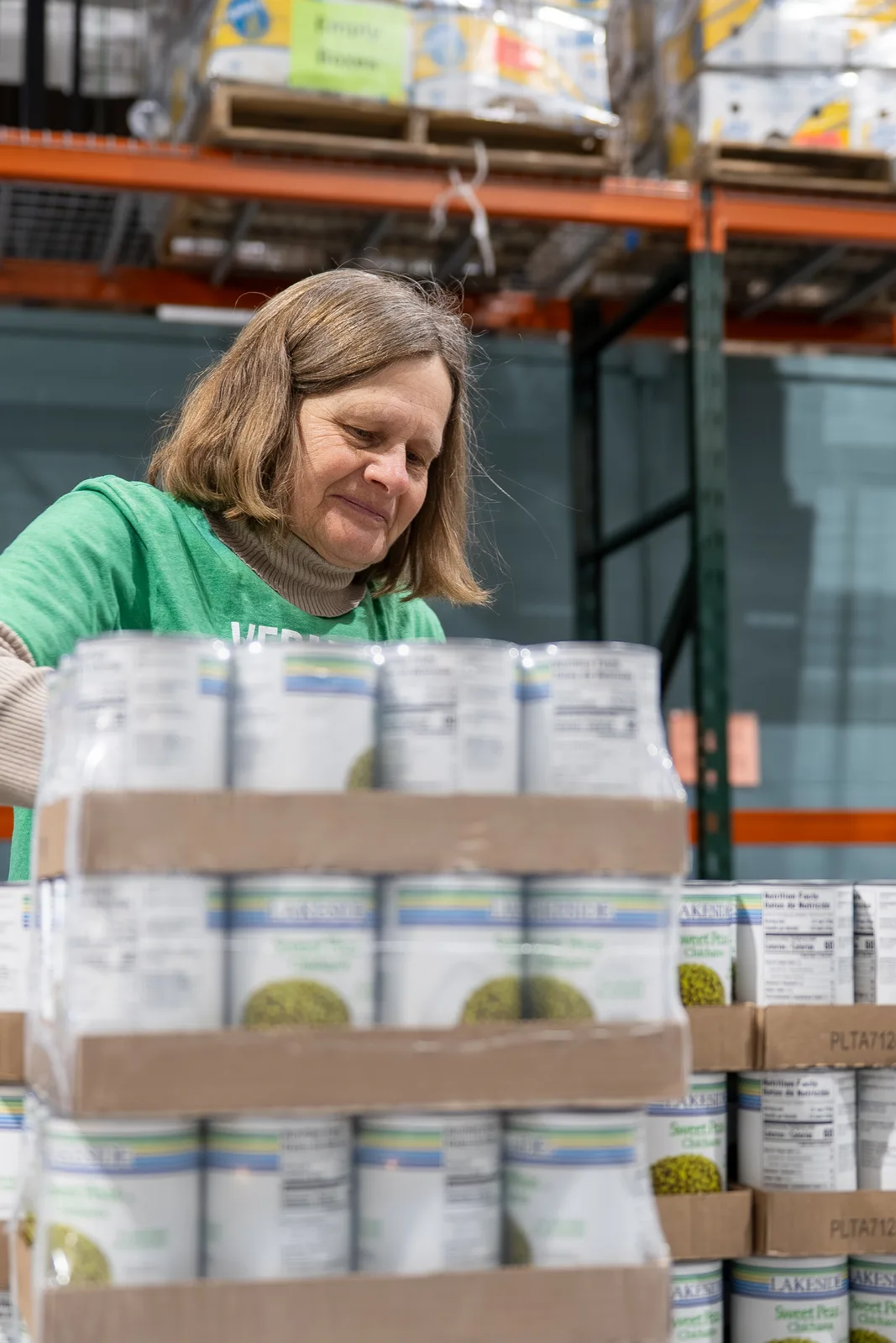 Person in green shirt arranging stacked boxes of canned goods in a warehouse