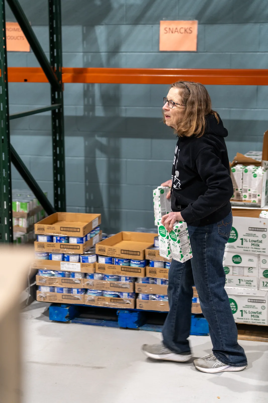 Person in black sweatshirt holding milk carton near stacked boxes in warehouse with metal shelving