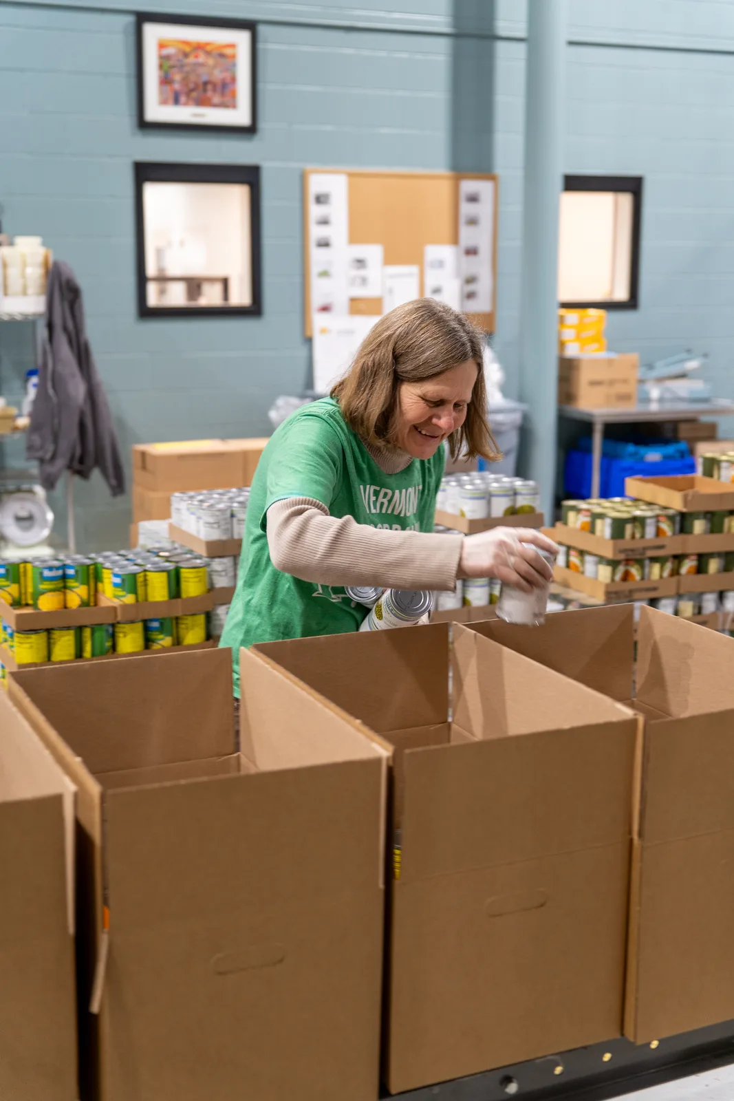 Person in green shirt placing canned food into cardboard box on table in warehouse