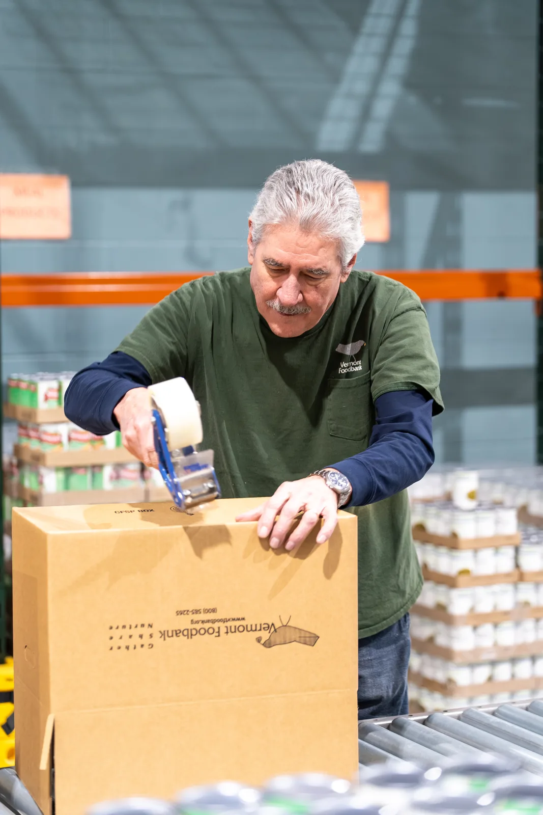Person in green shirt taping a Vermont Foodbank box on conveyor belt in warehouse