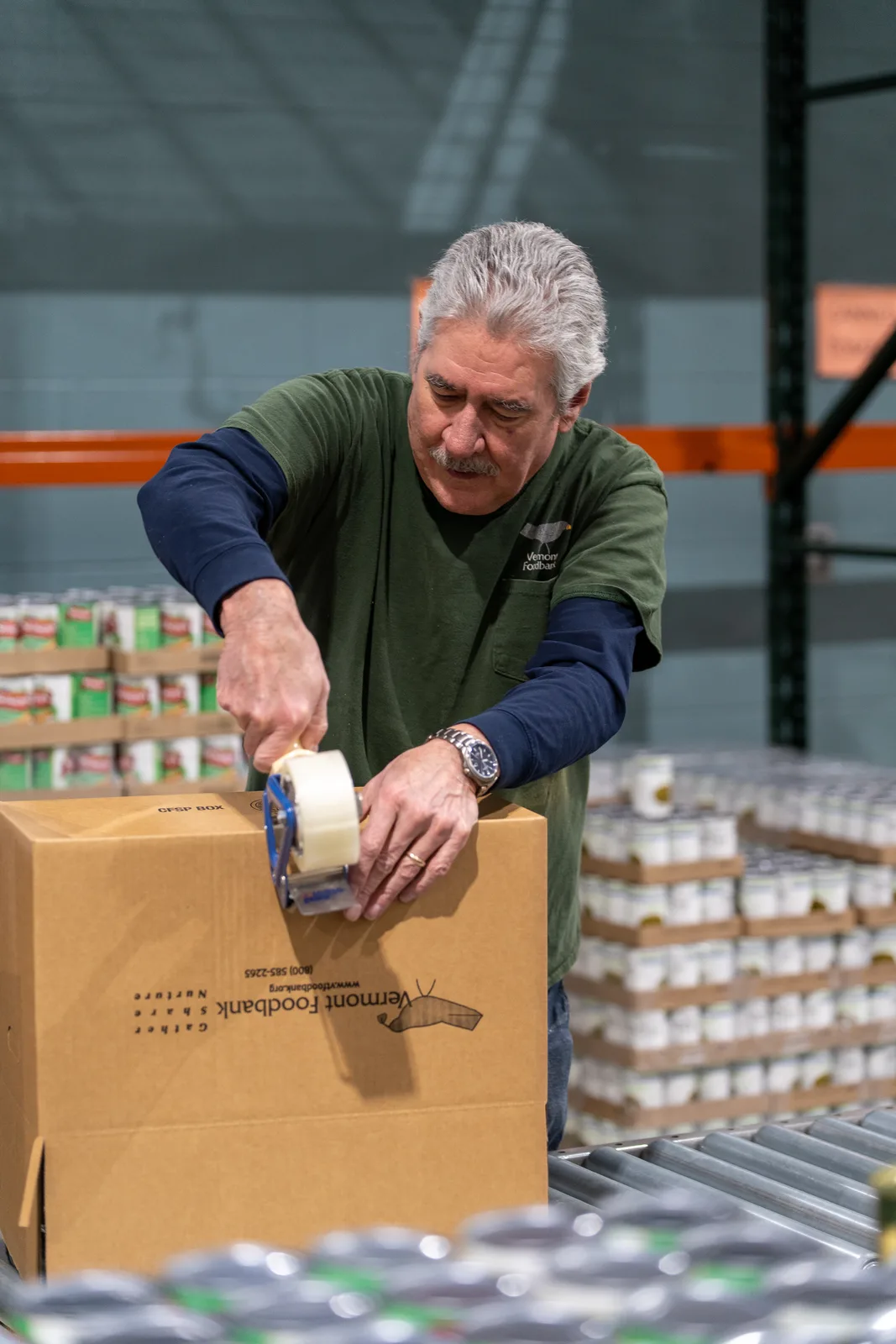 Person in green shirt taping a box on conveyor belt with stacked cans in background