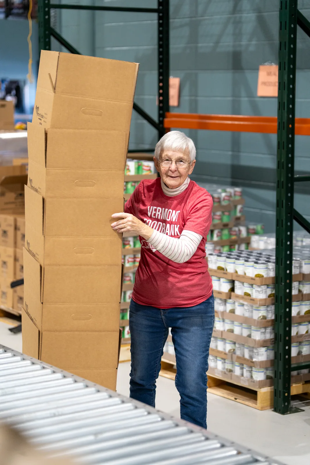 Person in red shirt holding stacked cardboard boxes near conveyor belt in warehouse