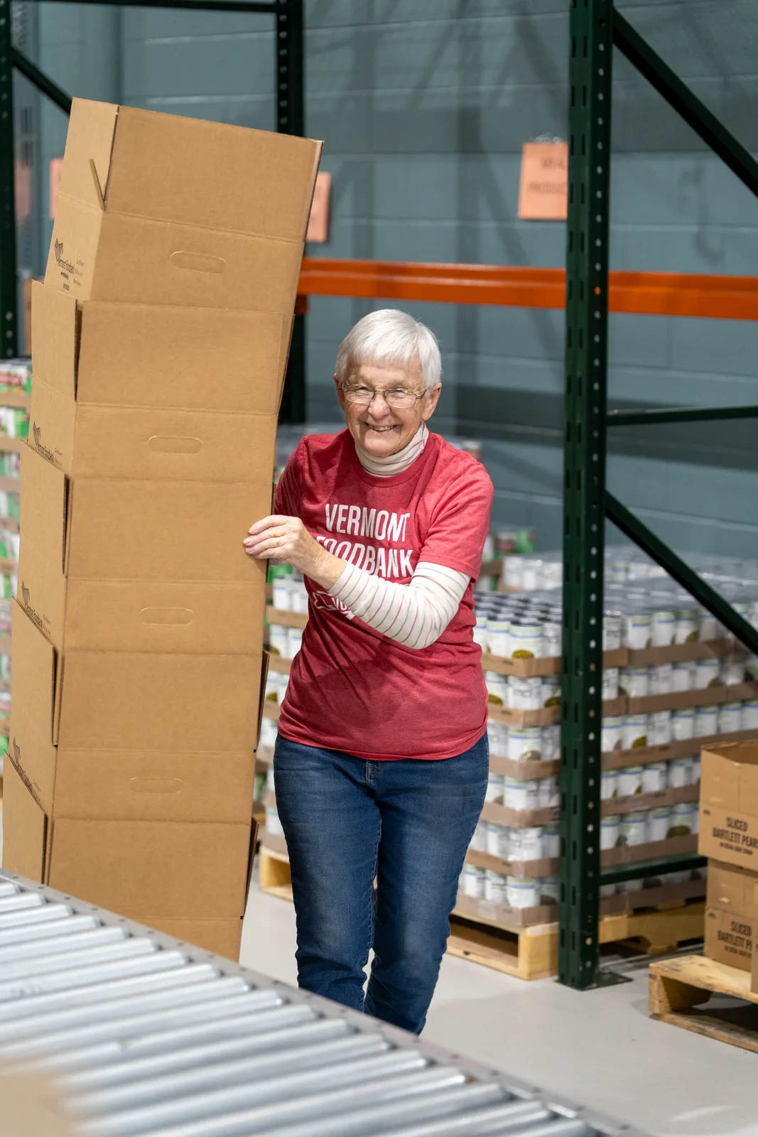 Person in red shirt holding stacked cardboard boxes near conveyor belt and shelves