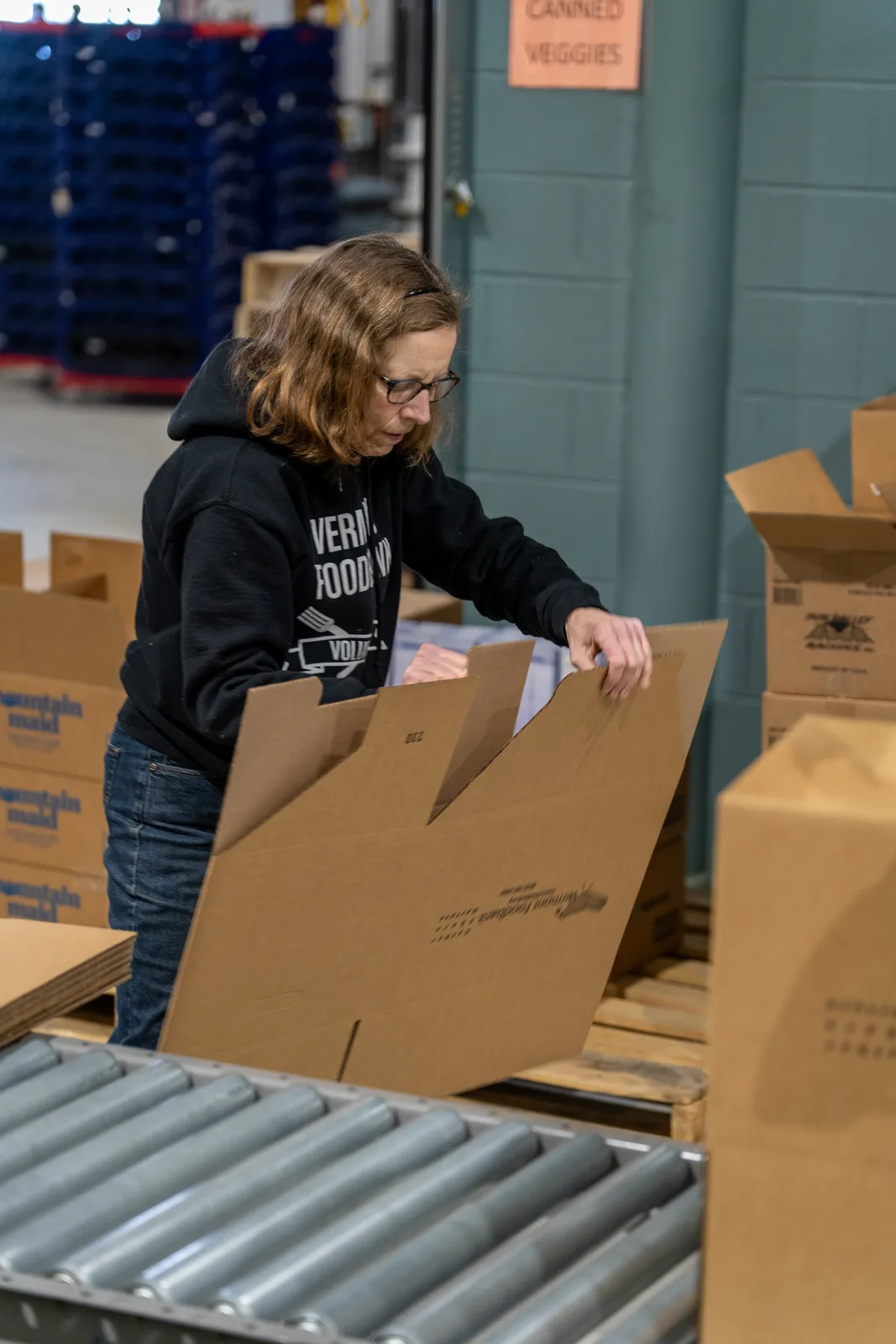 Person in black sweatshirt assembling a cardboard box on a conveyor in warehouse