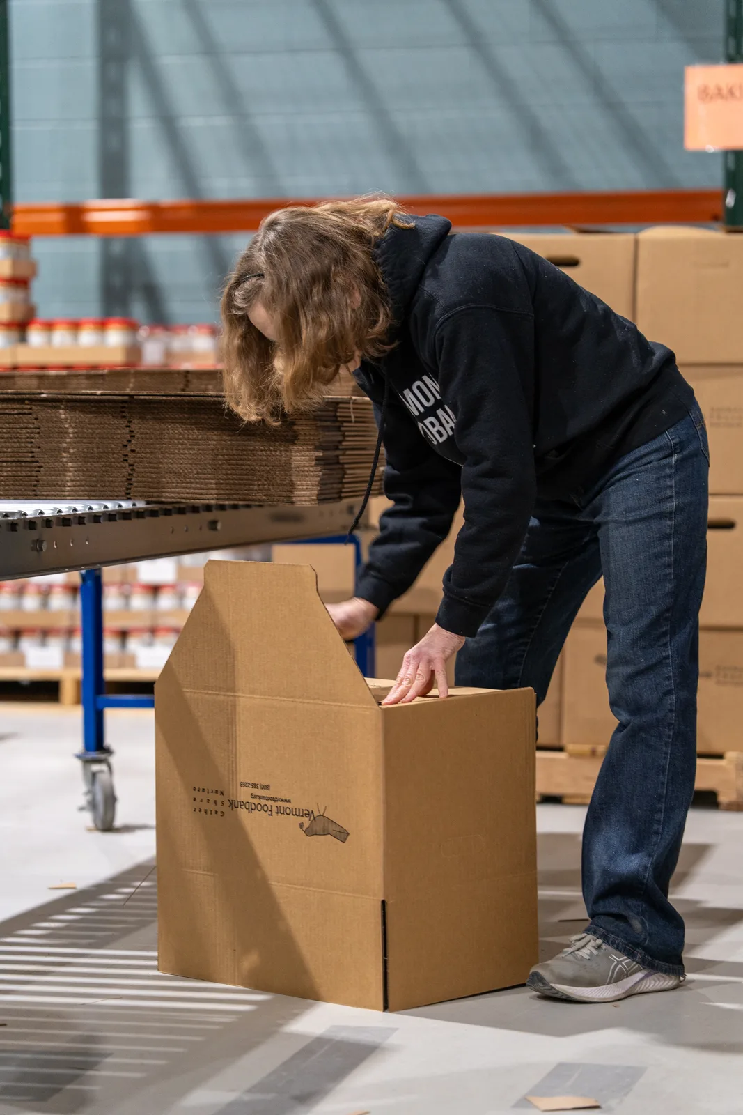 Person in black sweatshirt bending to adjust a cardboard box on conveyor