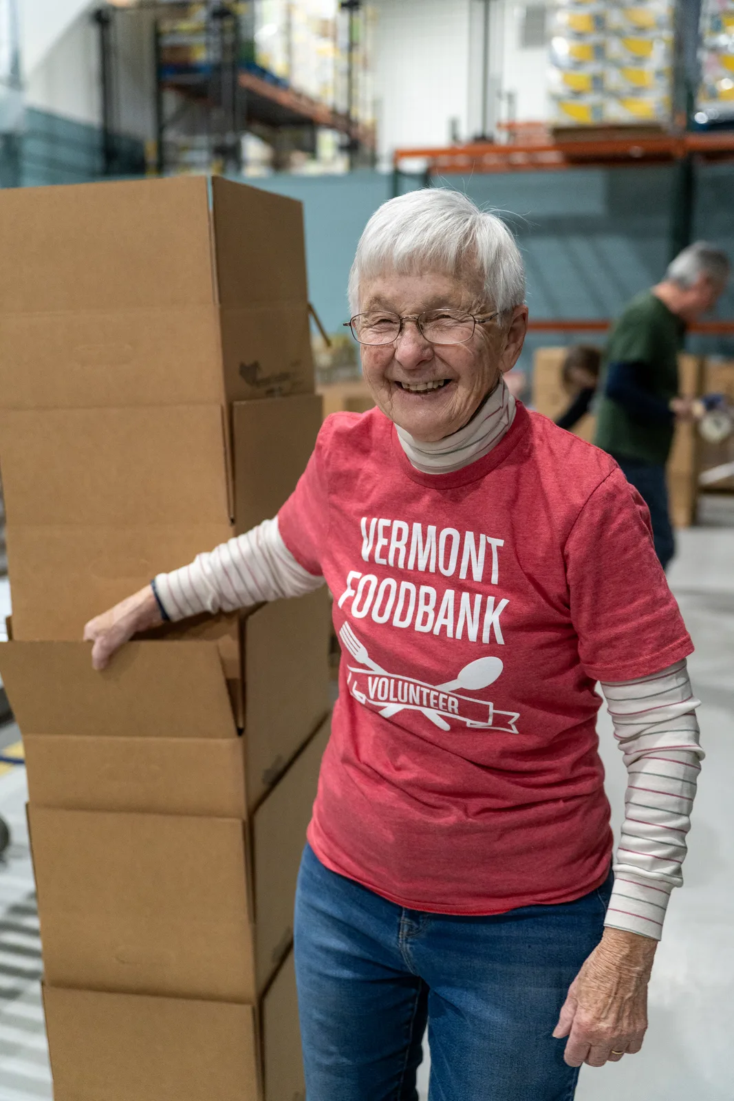 Person in red shirt standing beside tall stack of cardboard boxes in warehouse