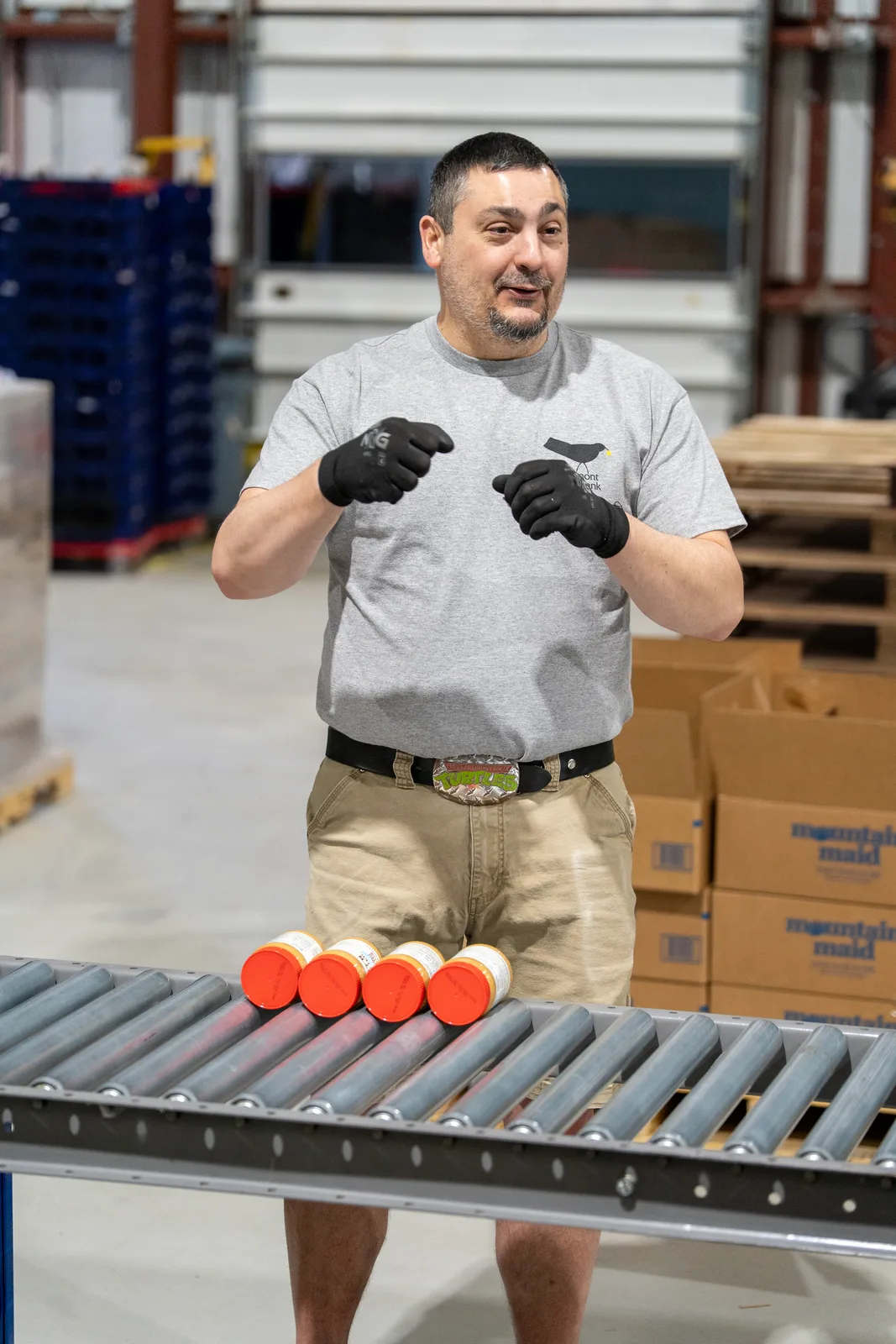 Person in gray shirt gesturing near conveyor with orange canned goods