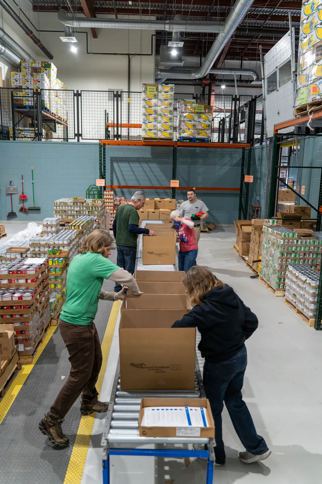 Four people packing cardboard boxes on a conveyor in a warehouse with shelves of food