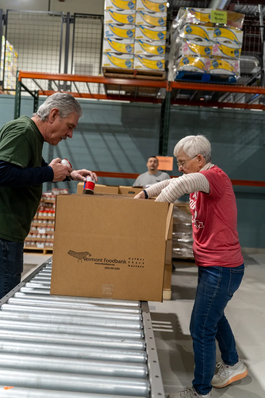 Two people taping a Vermont Foodbank box on a conveyor with shelves in background