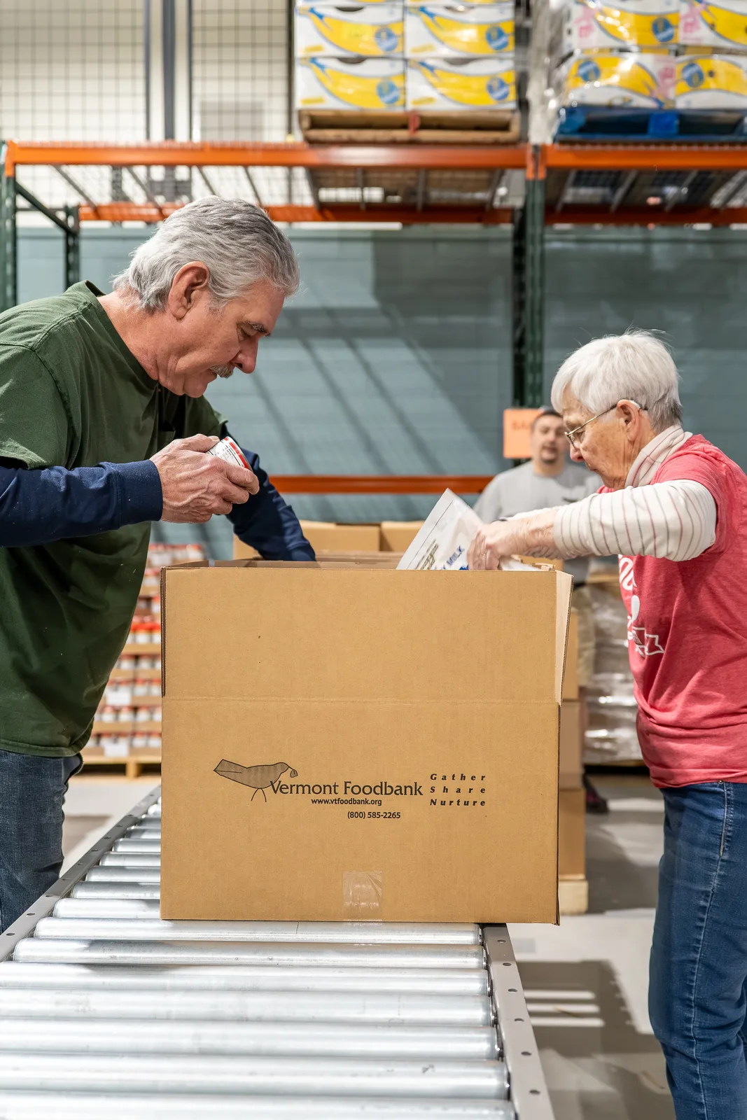 Two people taping a Vermont Foodbank box on a conveyor with shelves of food