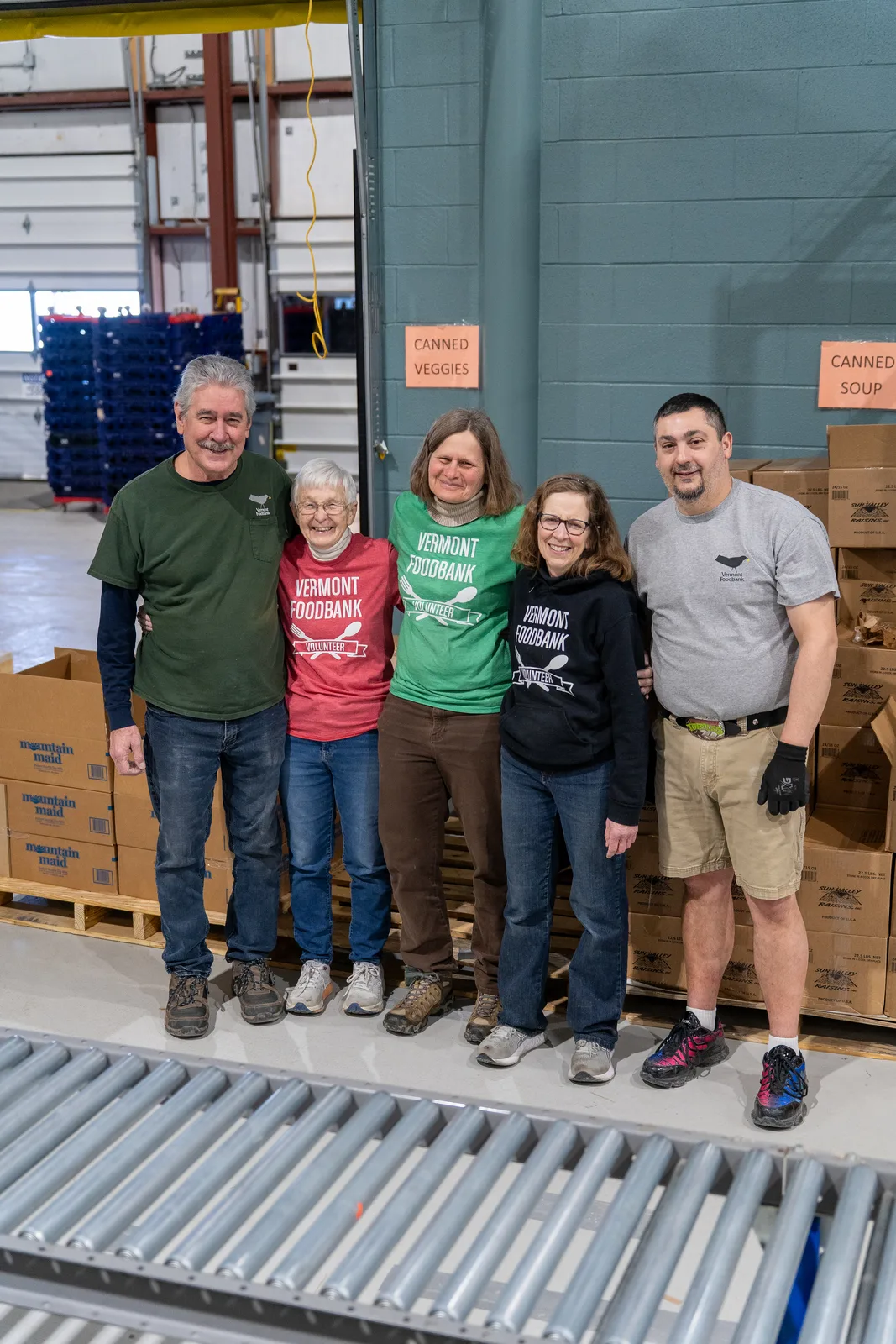 Five people standing in front of a conveyor with stacked boxes and warehouse shelves