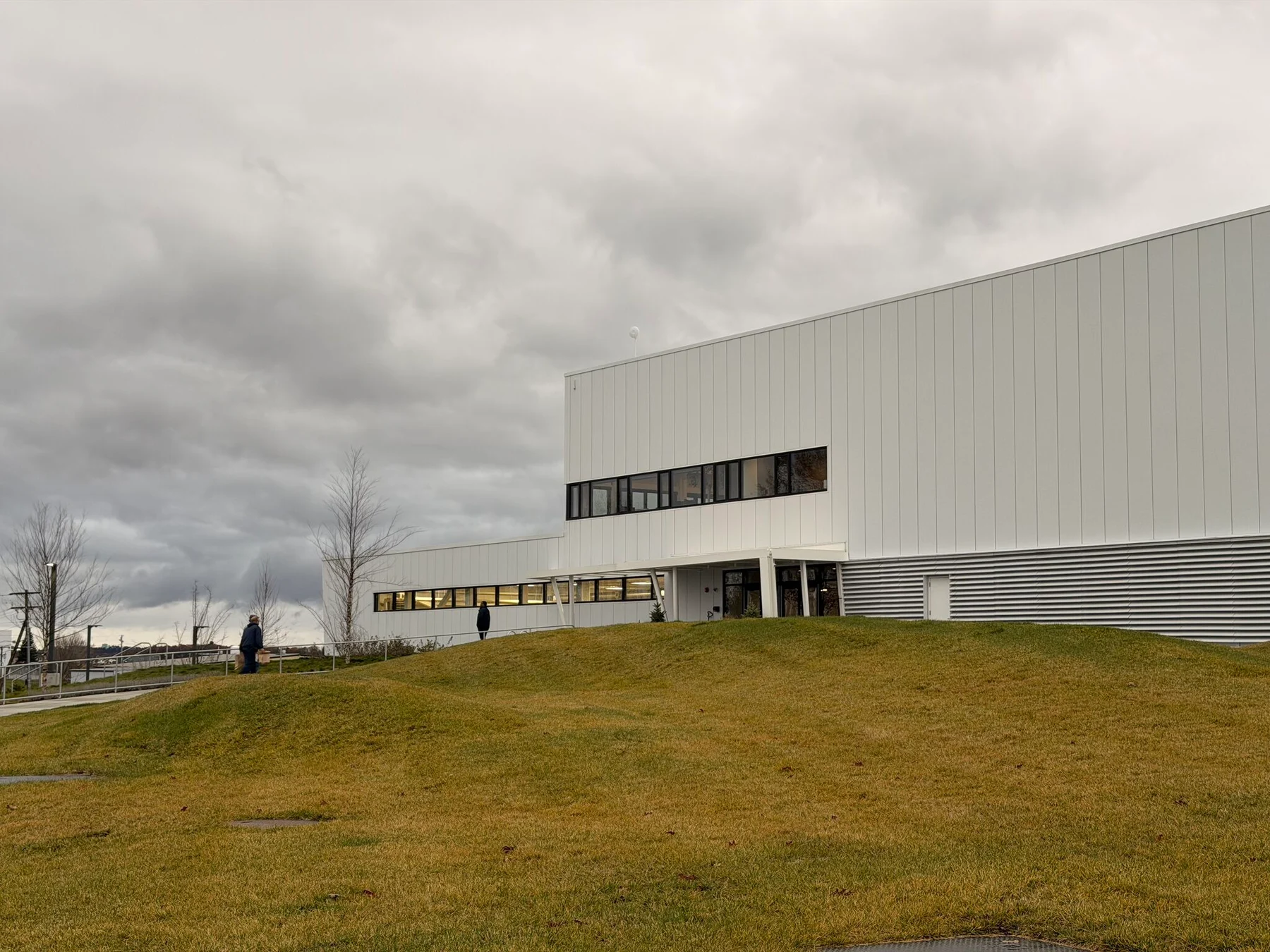A large white BETA Technologies facility stands on a grassy slope beneath dark clouds.