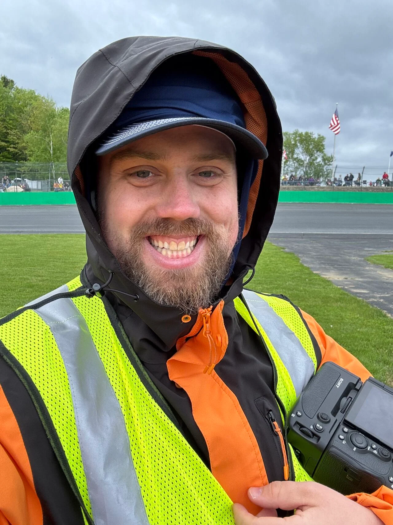 Oliver Ames takes a rainy racetrack selfie in an orange jacket and reflective safety vest while holding a camera.