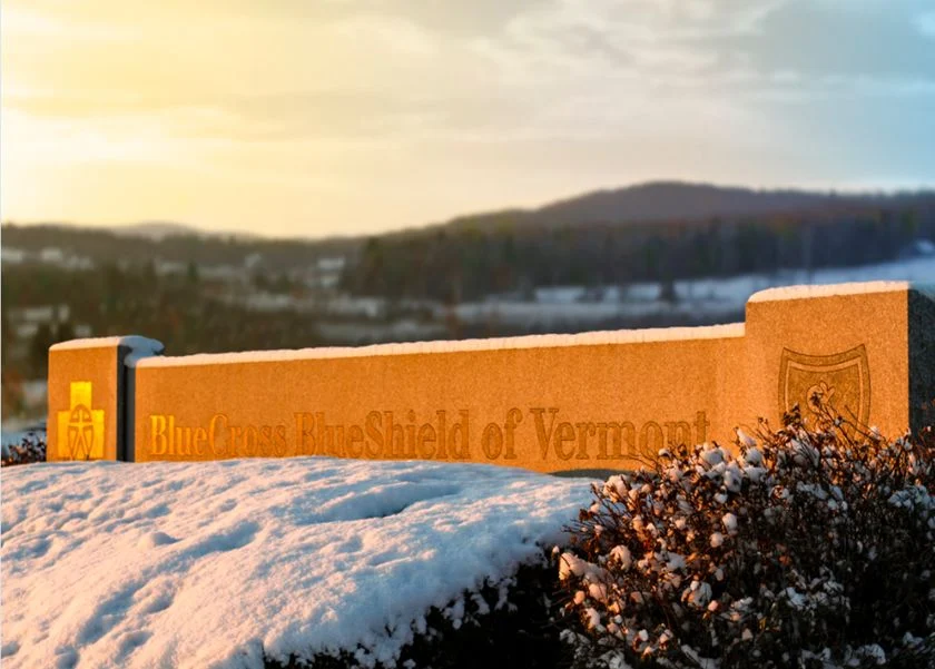 The Blue Cross and Blue Shield of Vermont sign glows in late-day sunlight above a snow-covered hedge.