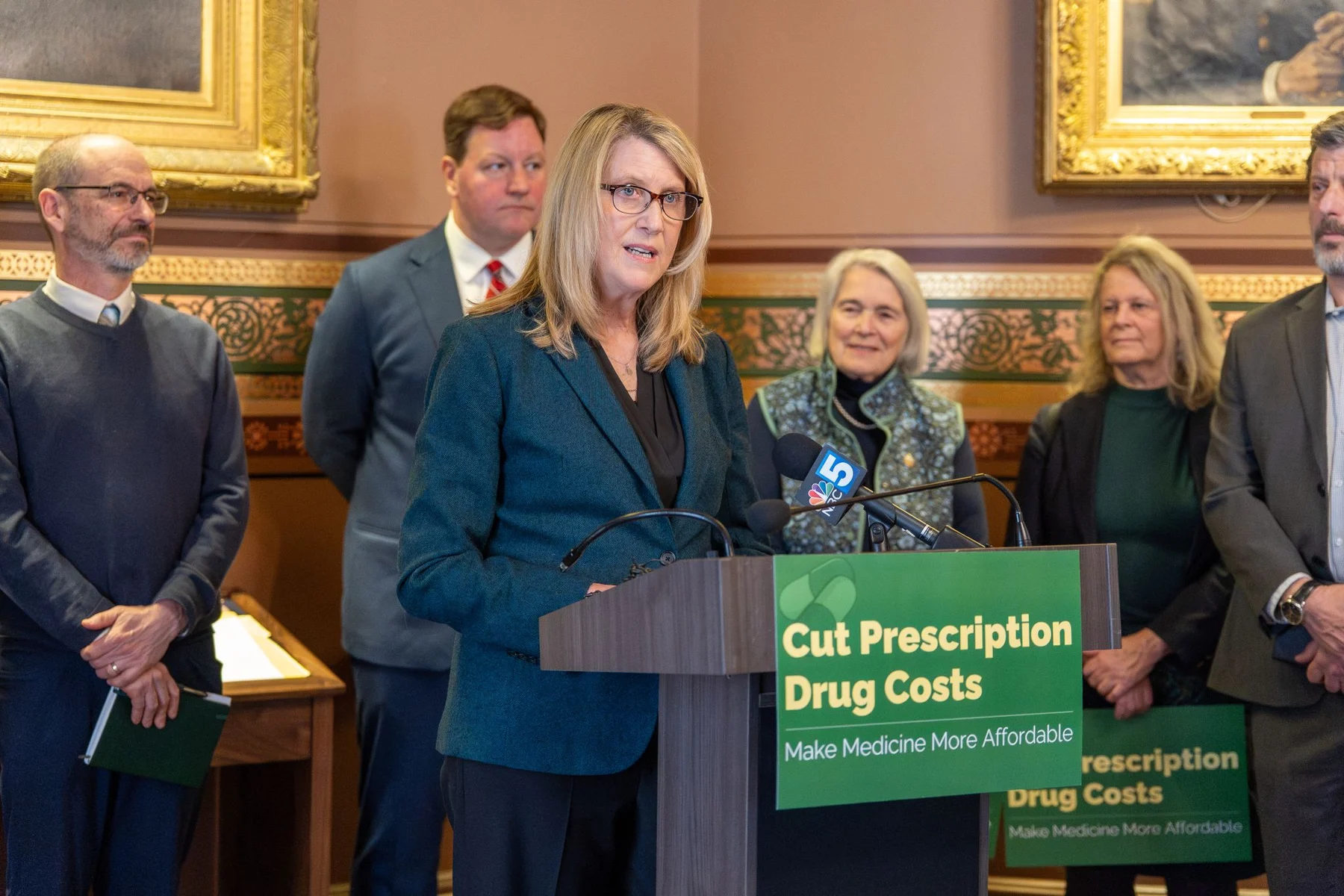 A woman speaks at a State House podium beside supporters and a sign calling for lower prescription drug costs.