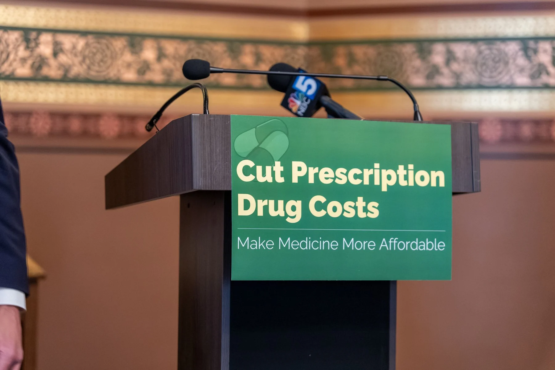 An empty State House podium displays a green sign reading ‘Cut Prescription Drug Costs: Make Medicine More Affordable.’
