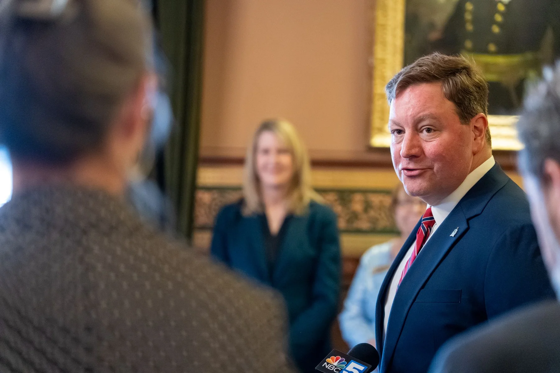 A man in a navy suit and red tie speaks to reporters at the State House.
