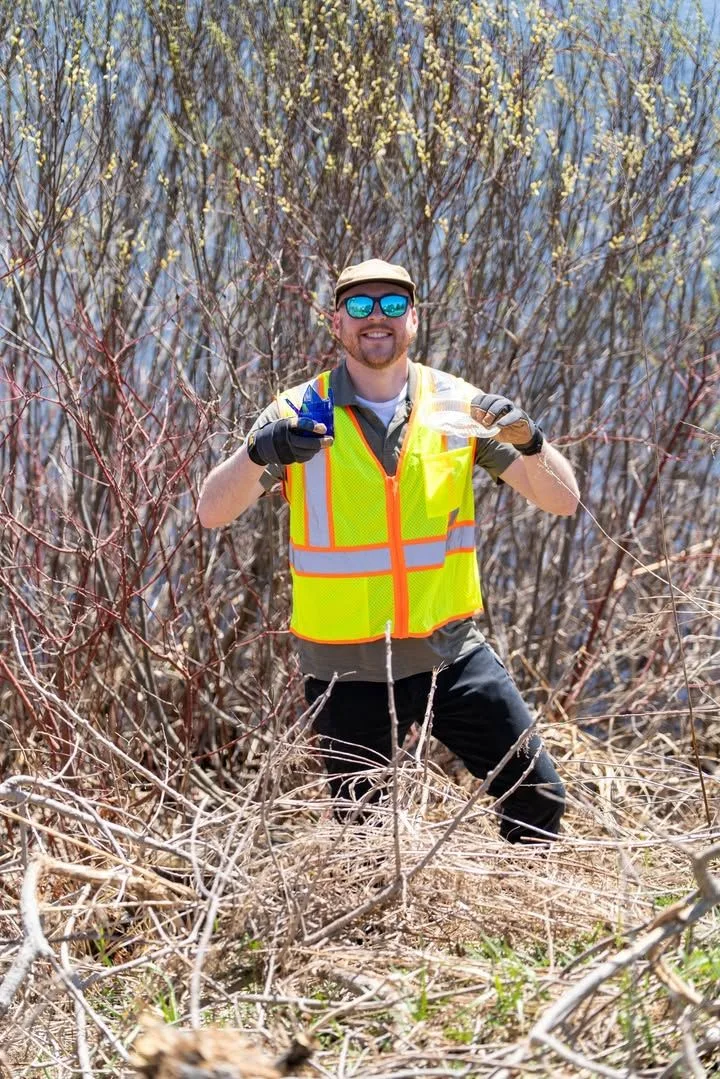 A volunteer in a safety vest works among roadside brush.
