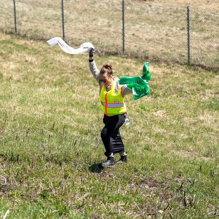 A volunteer lifts a green cleanup bag on a grassy hillside.